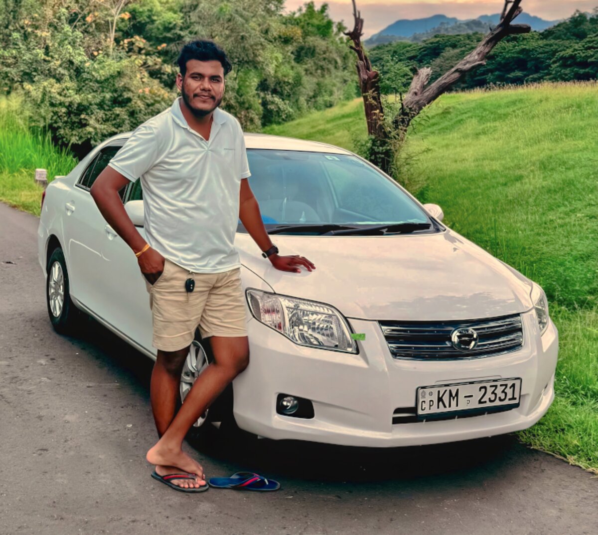 Experienced Kayeli Lanka Tours chauffeur standing beside a clean white sedan on a scenic Sri Lankan road